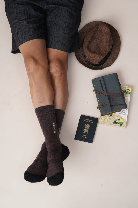 A person in dark shorts and brown socks lies on a neutral background beside a brown hat, a journal, a passport, and travel brochures, conveying a relaxed, travel-ready vibe.