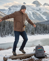 Man in Faux leather Jacket standing on a log with a backpack and ski poles in a snowy landscape with mountains.