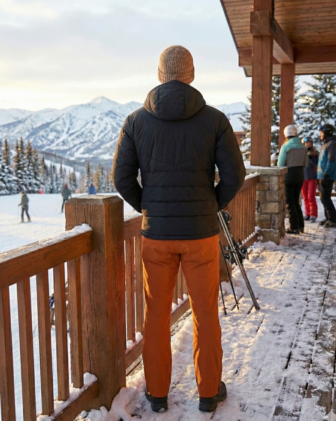 Person in winter clothing standing on a wooden deck with a snowy landscape and mountains in the background.