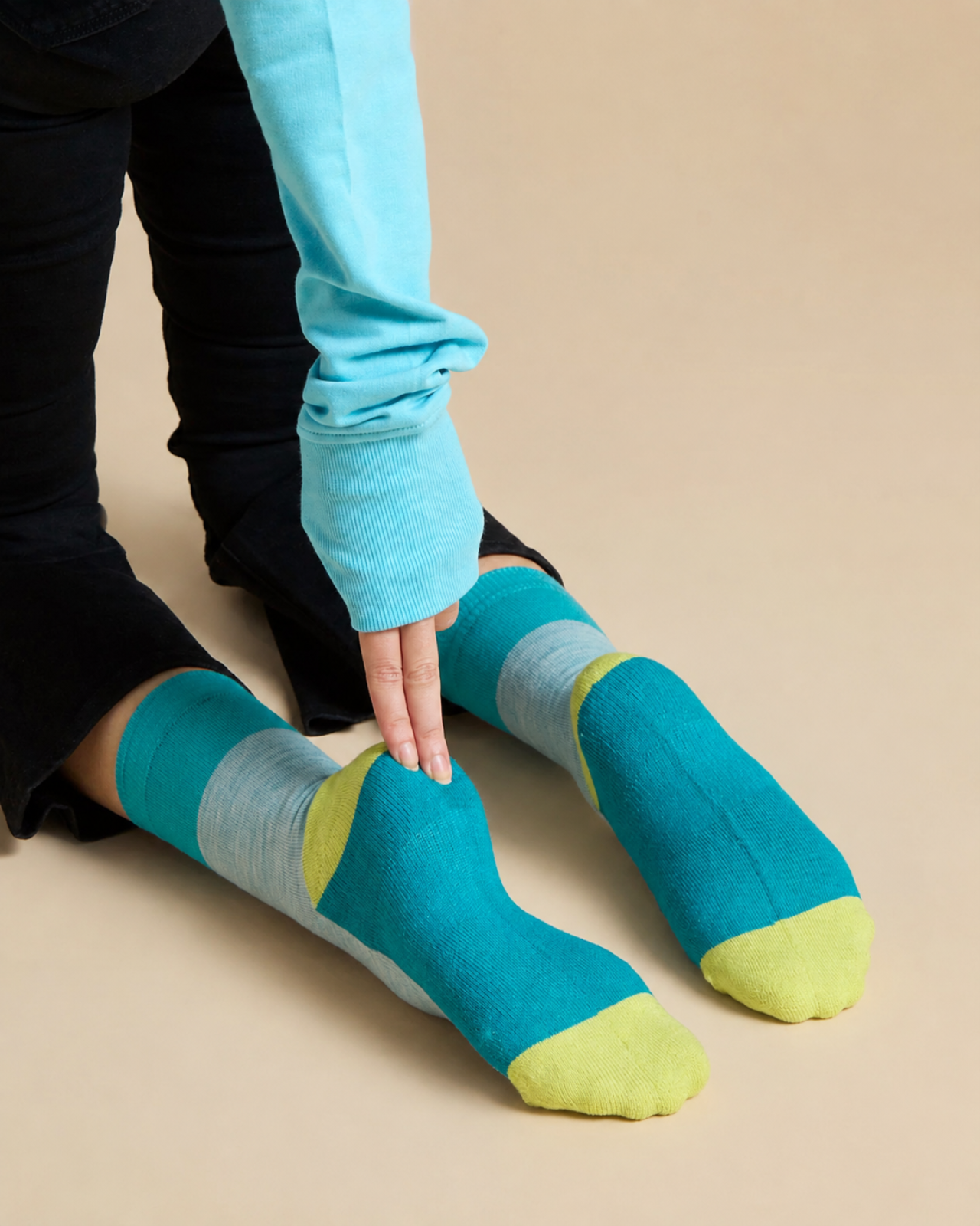 A person wearing colorful striped socks with a turquoise blue body, light gray bands, and lime green toes and heels, shown from the knees down against a plain beige background.
