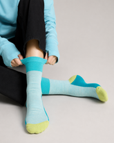 A person sitting on a light gray floor wearing light aqua crew length technical socks with bright turquoise blue ribbed cuffs and heels, lime green toes, and black pants.