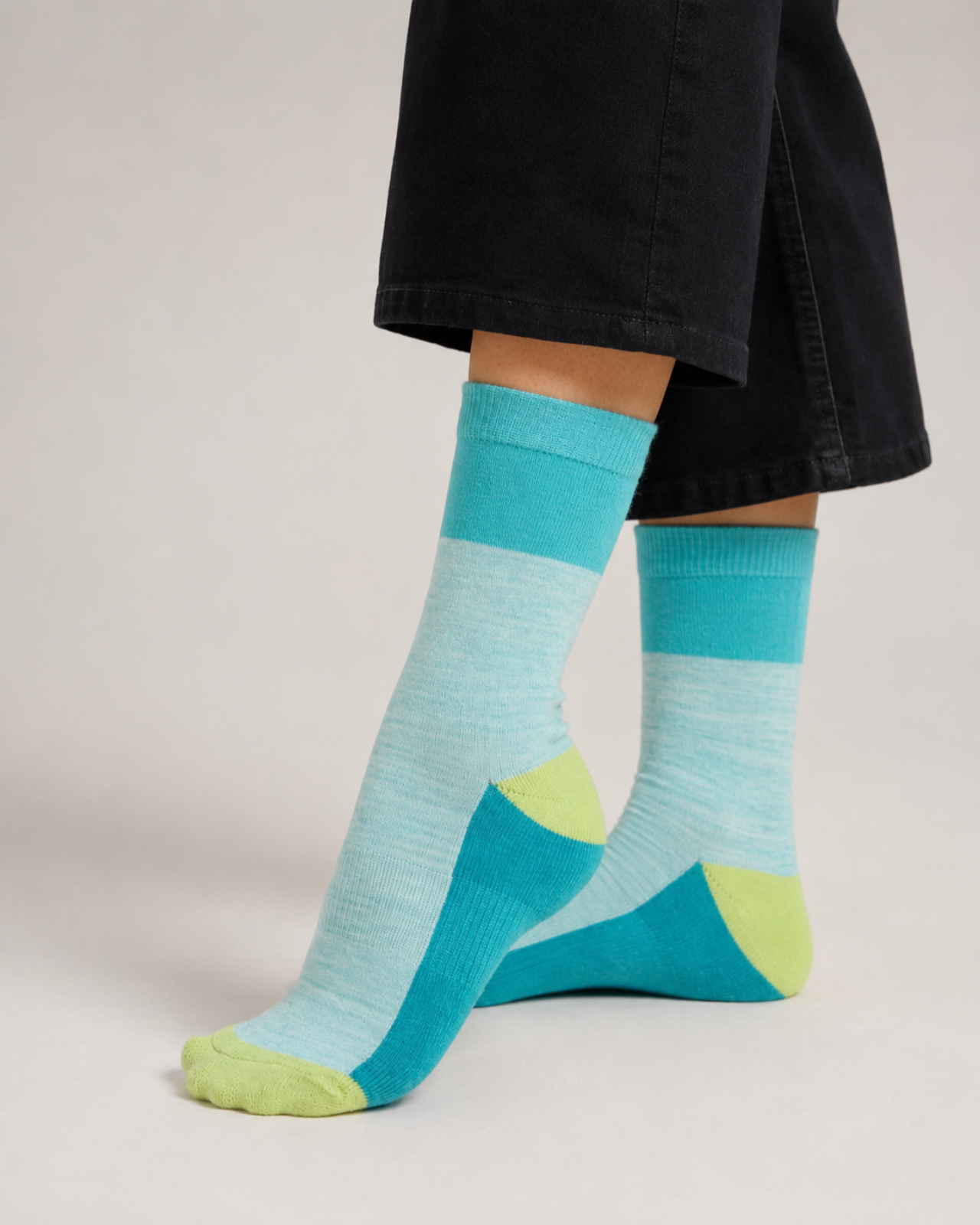 A pair of crew socks worn on a person's feet, featuring horizontal stripes of light blue and teal with lime green accents on the toes and heels, paired with black pants against a light gray studio background.