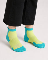 A close-up of a person's feet wearing low-cut athletic socks in neon green, paired with black pants, against a light gray studio background.