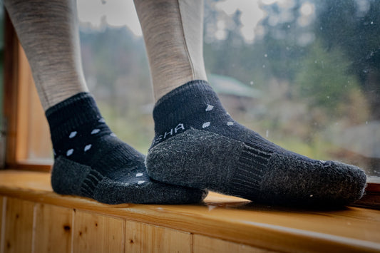 Close-up of feet in black, patterned woolen socks on a wooden windowsill. Background shows a blurred, green outdoor view, conveying a cozy atmosphere.