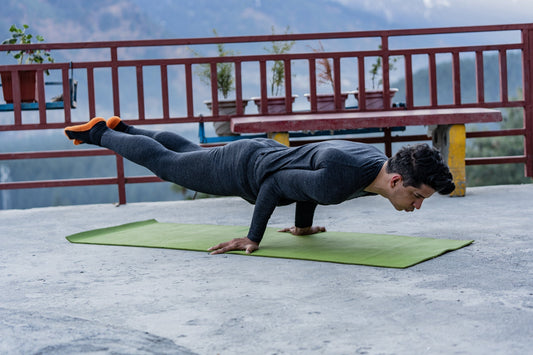 A person performs an advanced yoga plank on a green mat outdoors, surrounded by mountains and potted plants, conveying balance and focus.