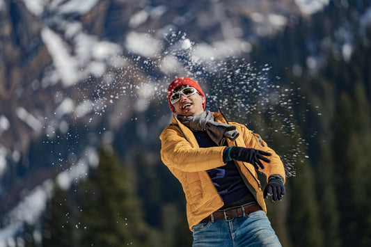 Man in a mustard winter jacket and red beanie playfully throwing snow, wearing gloves, scarf, and sunglasses in a snowy mountain setting.