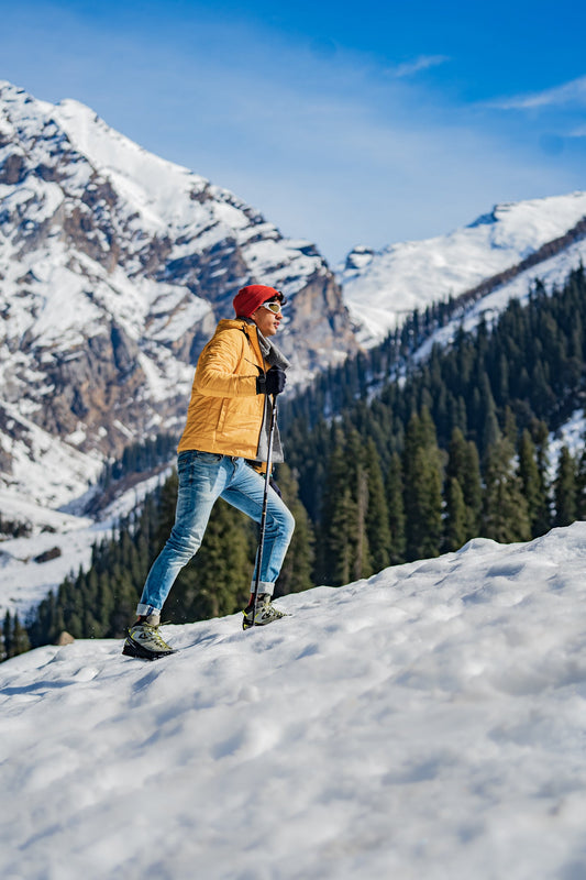 A person in a yellow jacket and red hat hikes up a snowy mountain slope with trekking poles, surrounded by snow-capped peaks and green pine trees under a clear blue sky.