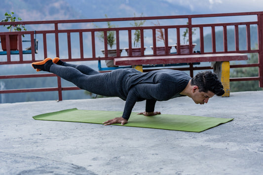 A person in a gray outfit performs an advanced yoga pose, balancing horizontally on a green mat outdoors. The background features a red fence and potted plants, with mountains in the distance, conveying strength and focus.
