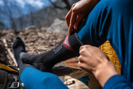 A person outdoors adjusts a black hiking sock with a red triangle pattern over blue leggings. The scene is calm and natural, with blurred trees in the background.