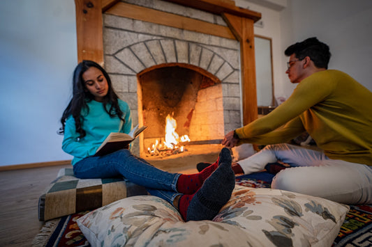 A woman reads a book by a cozy fireplace, wearing a turquoise sweater and jeans, while a man in a mustard sweater and white pants sits nearby.
