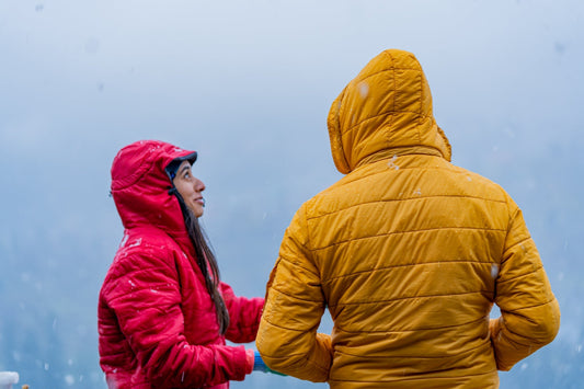 Two individuals stand in a snowy landscape, one in a vibrant red jacket and the other in a bright yellow one, against a foggy backdrop.