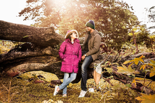 A couple playfully balances on a railway track surrounded by lush greenery. The woman in a pink coat and man in a green jacket exude joy and warmth.