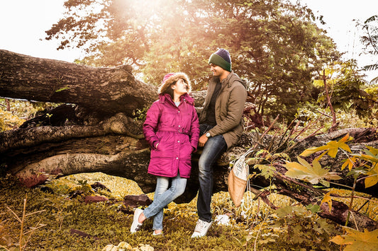 A couple sits on a large fallen tree trunk, surrounded by lush greenery and soft sunlight filtering through trees.