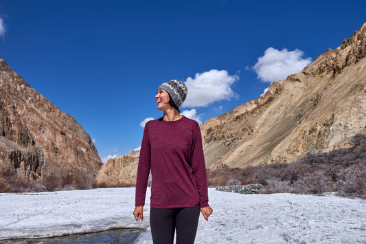 A person in a maroon sweater and gray beanie smiles broadly, standing on a snowy landscape with tall rocky mountains under a bright blue sky.