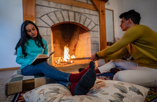 A man and a woman sitting near a lit fireplace, both wearing cozy Merino wool socks.