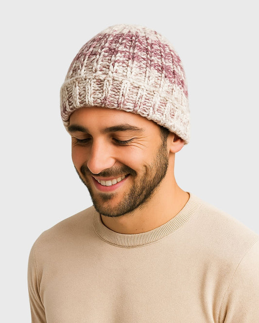 A smiling person wearing a knitted pink and cream beanie and a beige sweater against a plain gray background. The tone is warm and casual.