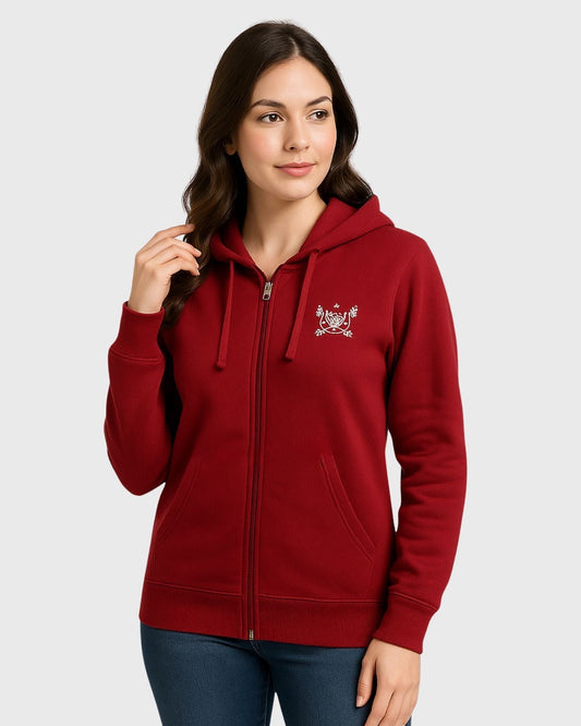 A woman in a red zip-up hoodie with a small white emblem on the chest stands against a light gray background. She has long, dark hair and wears jeans.