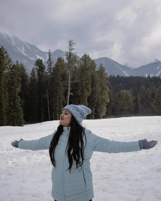 Woman wearing a sky-blue winter jacket, knitted beanie, and gloves, standing in the snow with arms outstretched, surrounded by pine trees and snow-covered mountains.