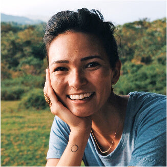 A woman with dark hair smiles warmly while resting her chin on her hand. She wears a blue shirt and a simple necklace. The background is a lush, green landscape.