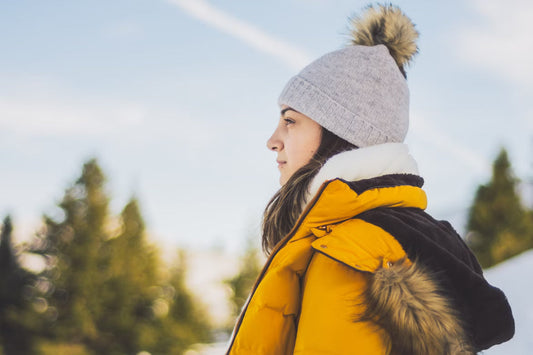 Profile of a person in a gray beanie and yellow winter coat with fur trim, standing outdoors. Background: blurred trees and a blue sky. Calm mood.