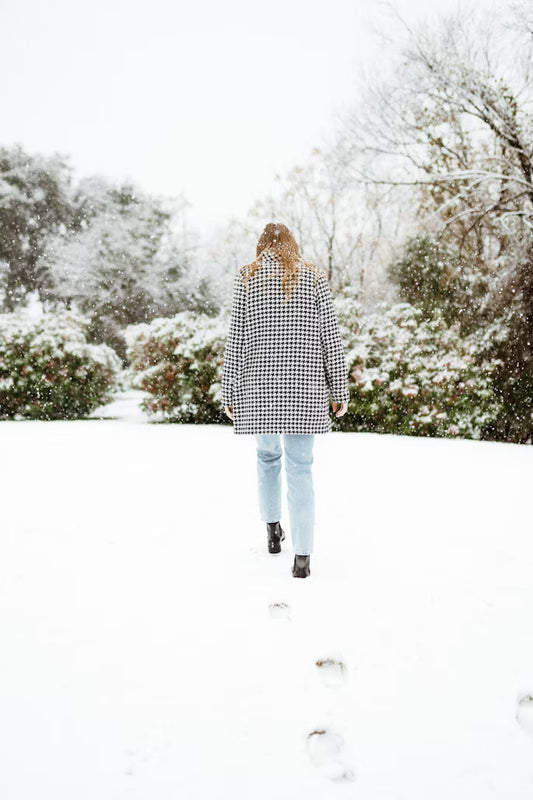A person in a houndstooth coat walks away in a snowy landscape. Snowflakes fall gently, footprints trail behind, and trees surround the scene.