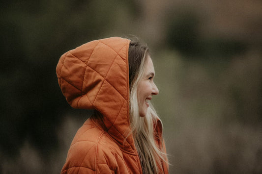 A woman in profile wearing an orange quilted hoodie stands outside, smiling. The background is blurred greenery, creating a warm and serene tone.