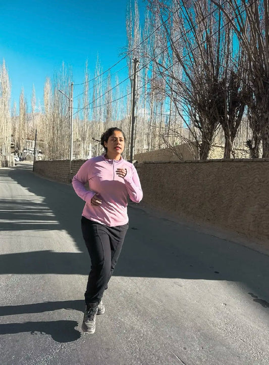A woman running through a scenic mountain trail surrounded by nature, wearing Kosha's lavender zipper jacket.
