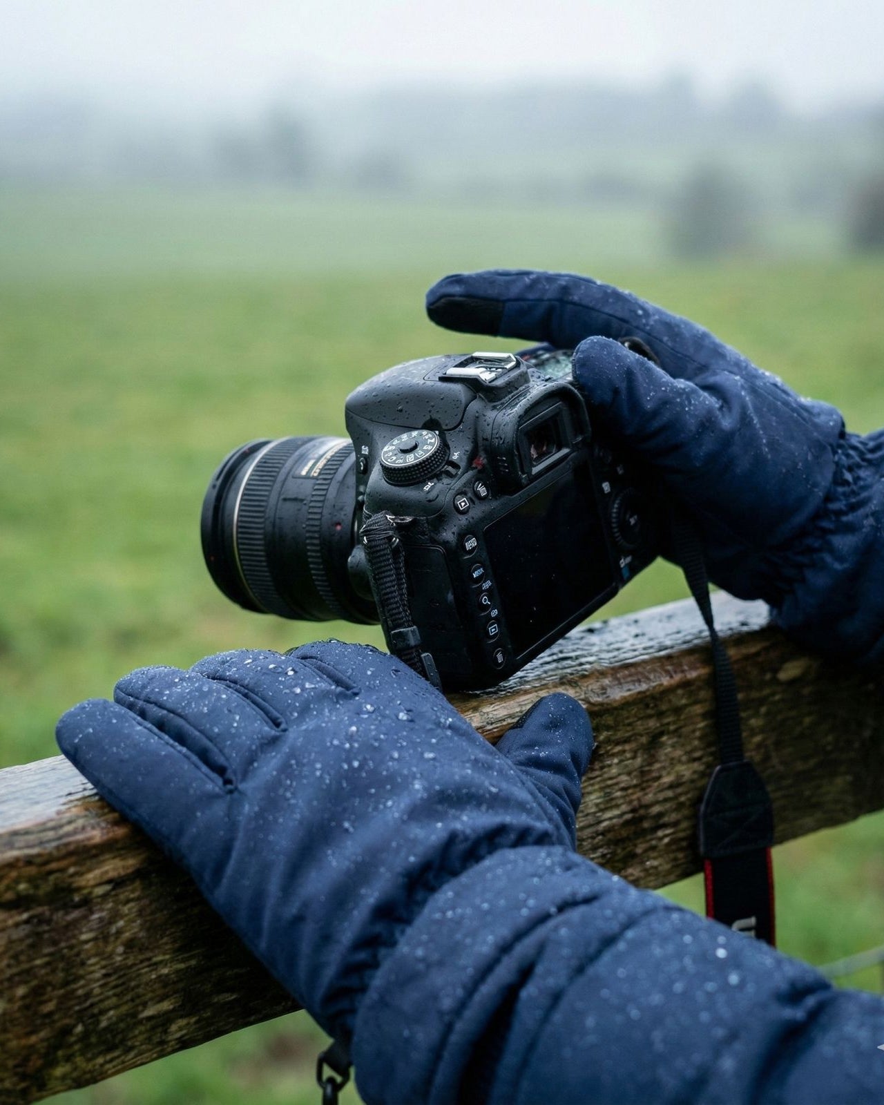 Person wearing Navy waterproof gloves holding a camera on a wooden fence with a blurred green field background
