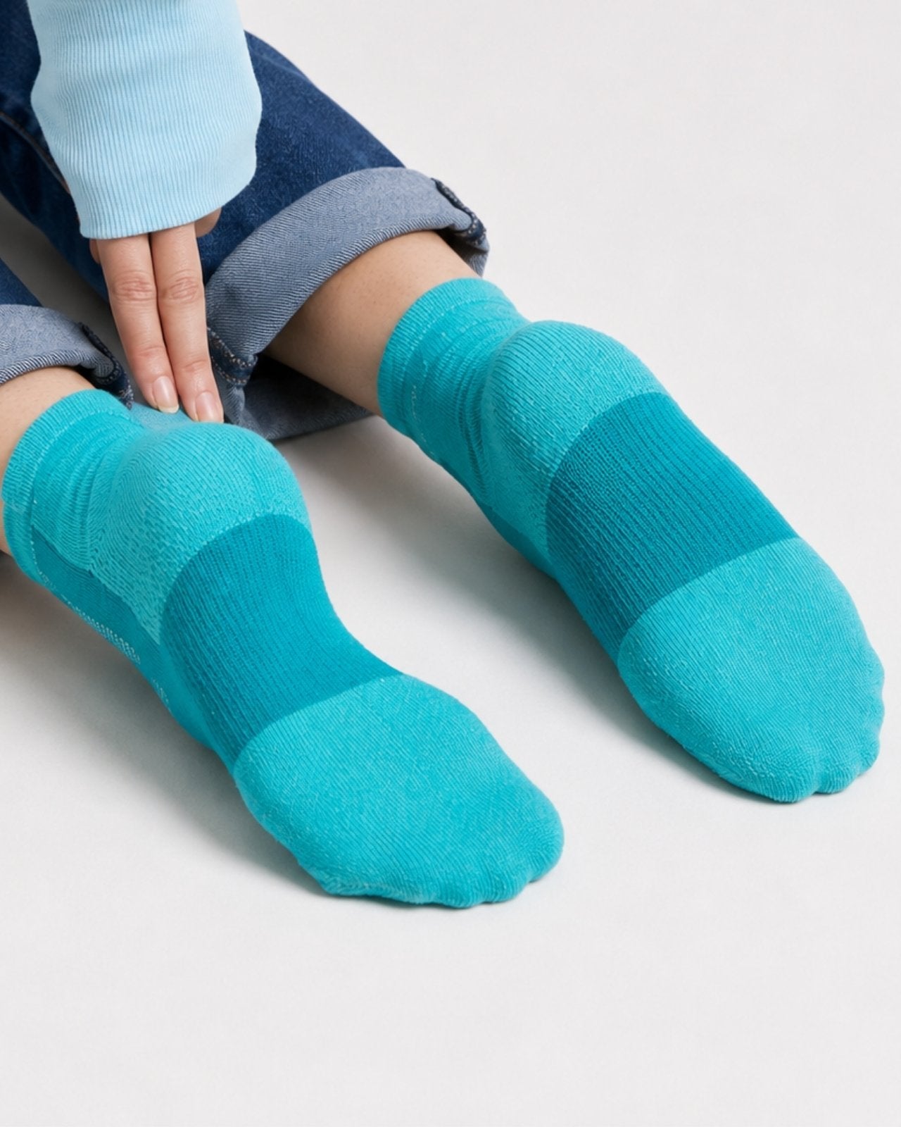 Wearing teal and blue socks with rolled-up blue jeans and a light blue long-sleeve top, set against a plain light gray background.